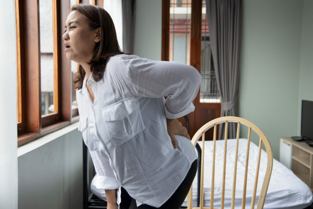 Woman in white shirt bending forward with hand on lower back, experiencing pain while sitting on bed