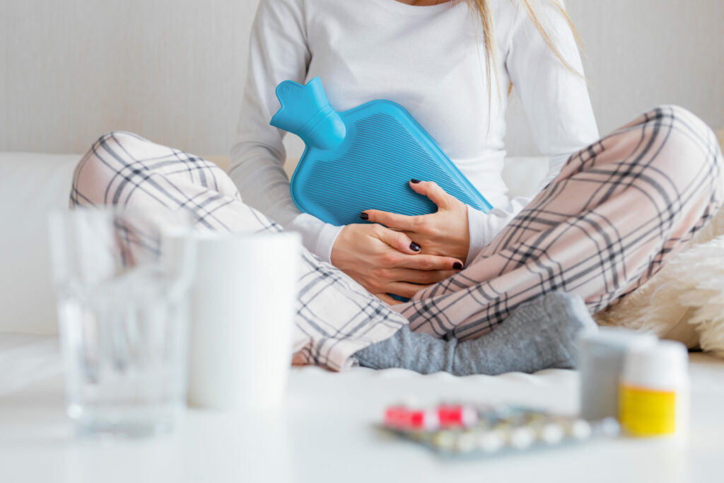 Person in pajamas holding blue hot water bottle for pain relief, with medications and glass of water nearby.