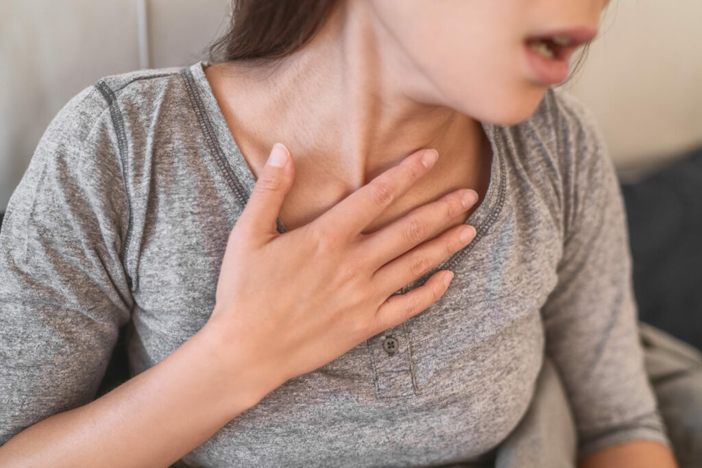 Woman in gray shirt holding her chest with hand, appearing to experience discomfort or pain