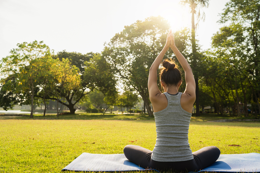 Woman sitting in yoga pose with hands joined above head in a park with green grass and trees