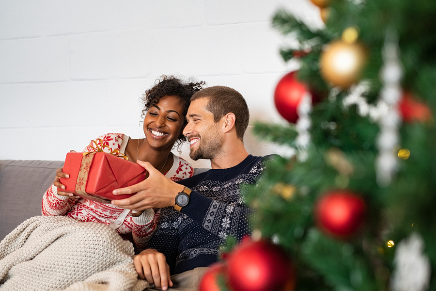 Smiling couple on couch exchanging wrapped red gift with decorated Christmas tree in background