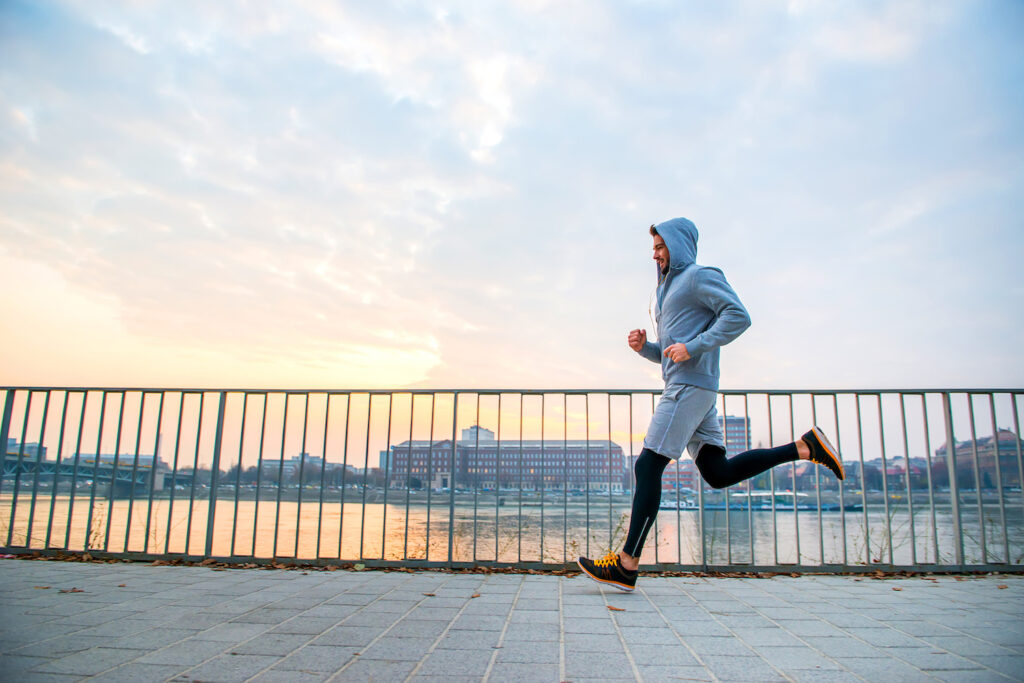 Runner in blue hoodie jogging on waterfront path with metal railing and city skyline behind