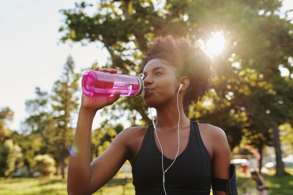 Woman in black sports top drinking from pink water bottle while wearing earbuds in sunny park with trees