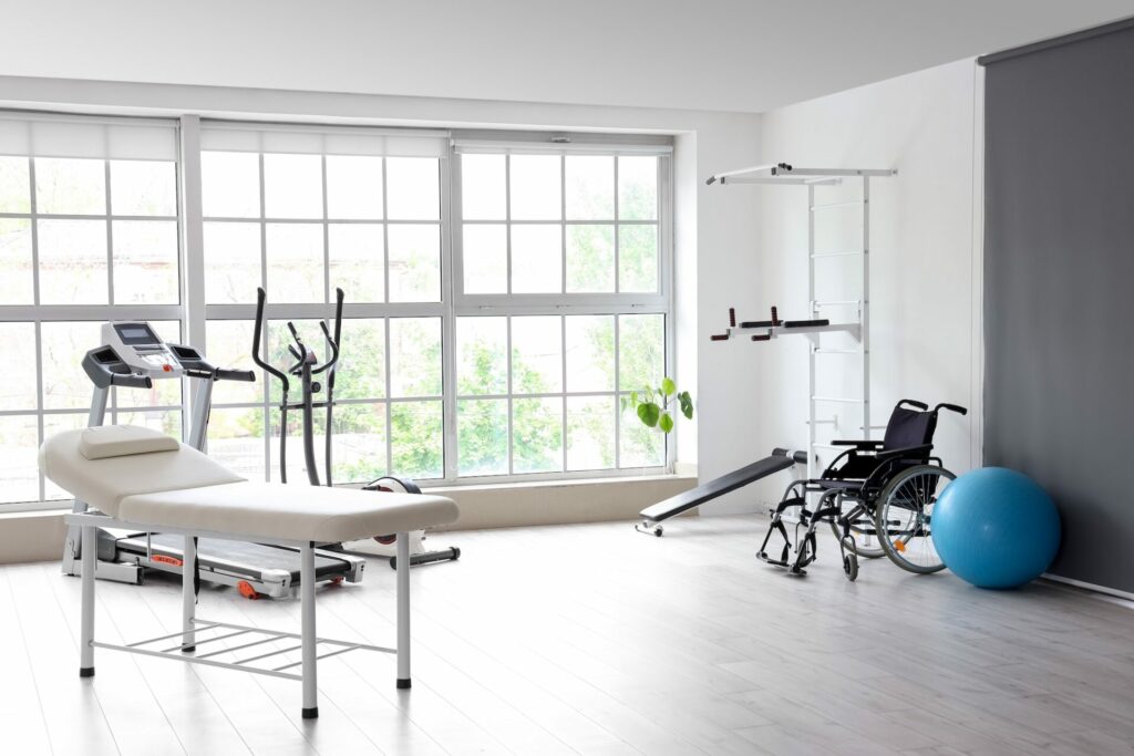 Modern rehabilitation clinic with medical equipment including treadmill, stationary bike, treatment table, wheelchair, exercise ball, and large windows overlooking trees.